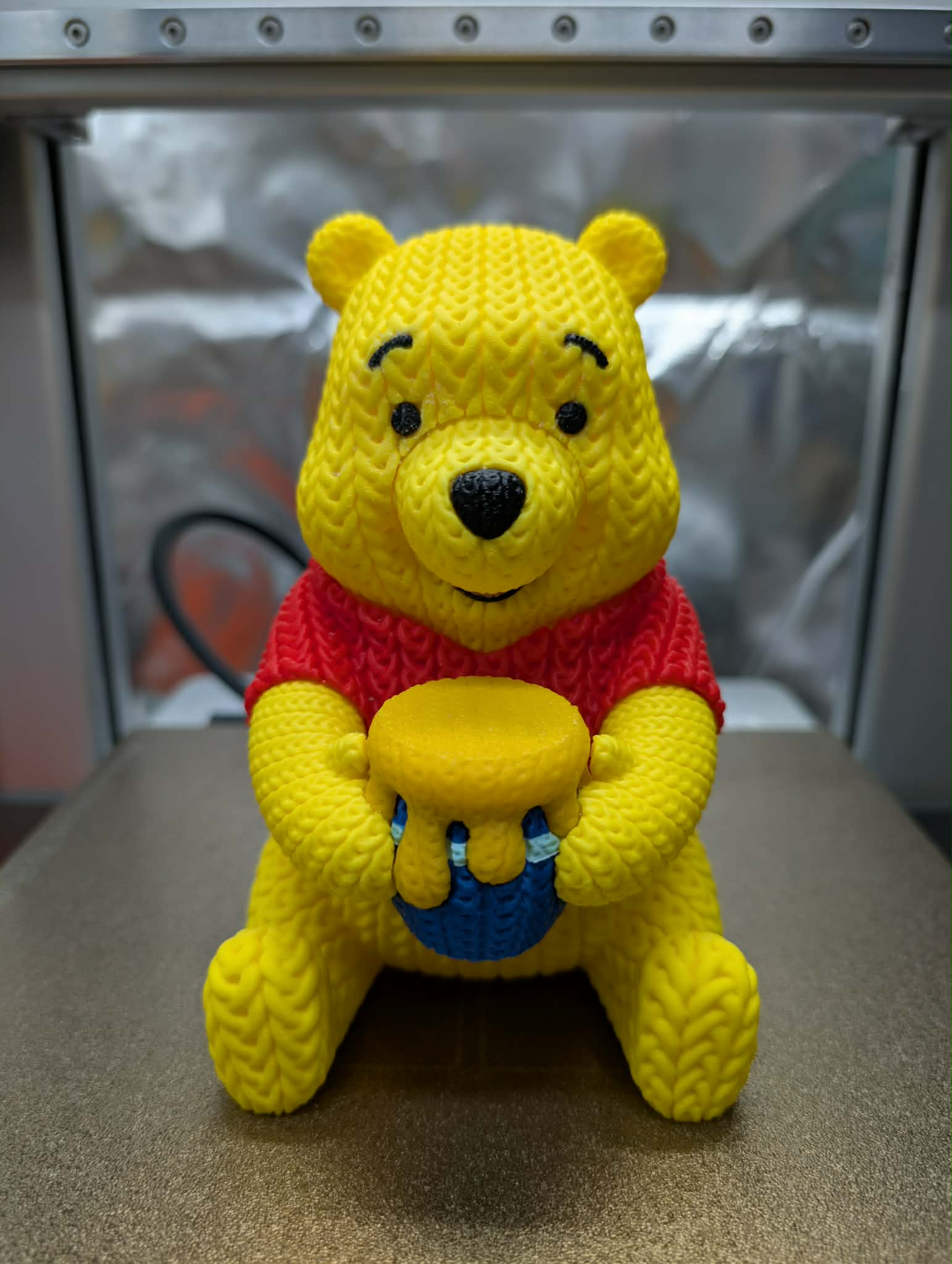 Yellow teddy bear plush toy with a red sweater and blue ball on a metallic surface.
