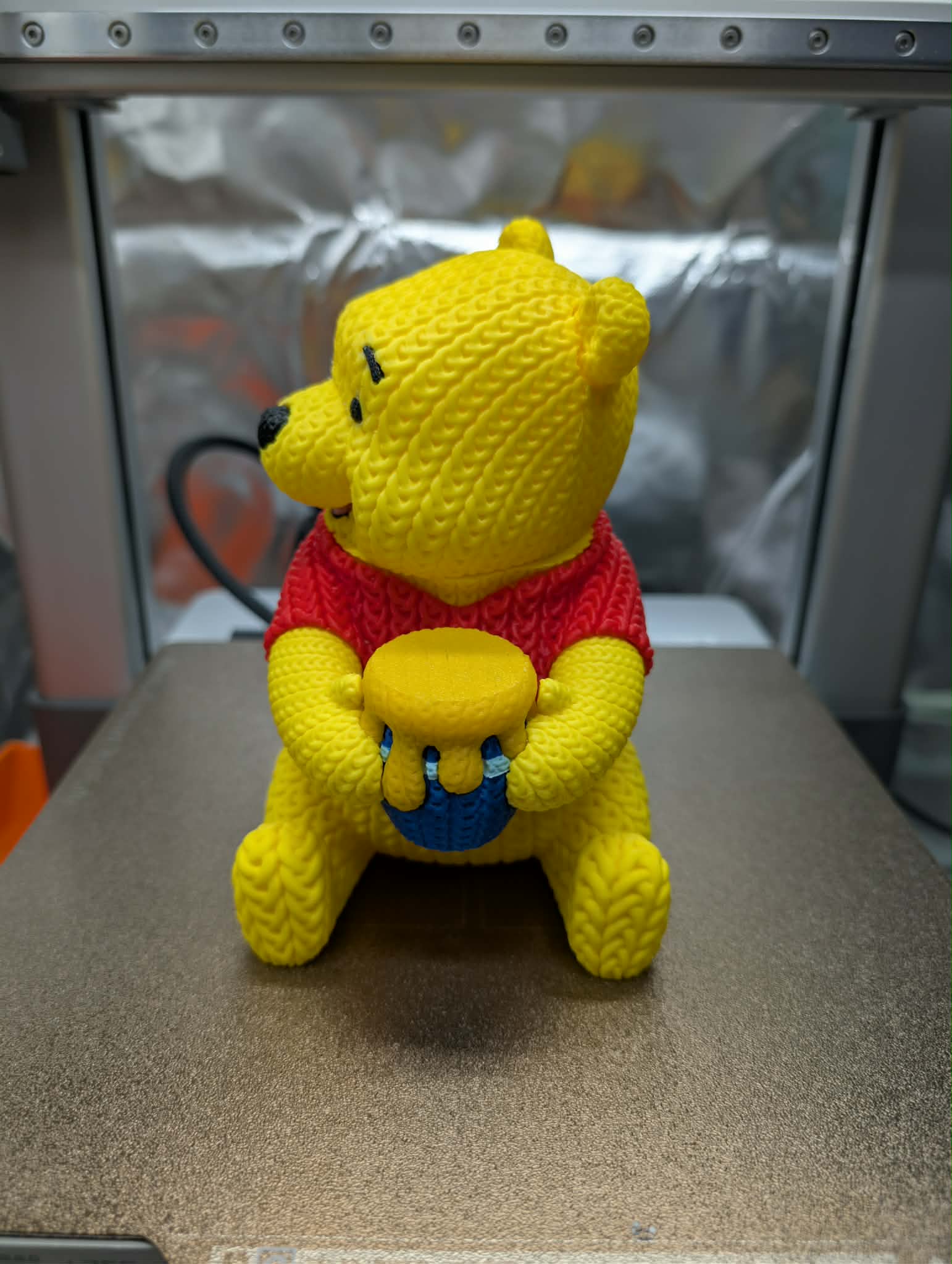 Yellow teddy bear with a red sweater on a metallic surface