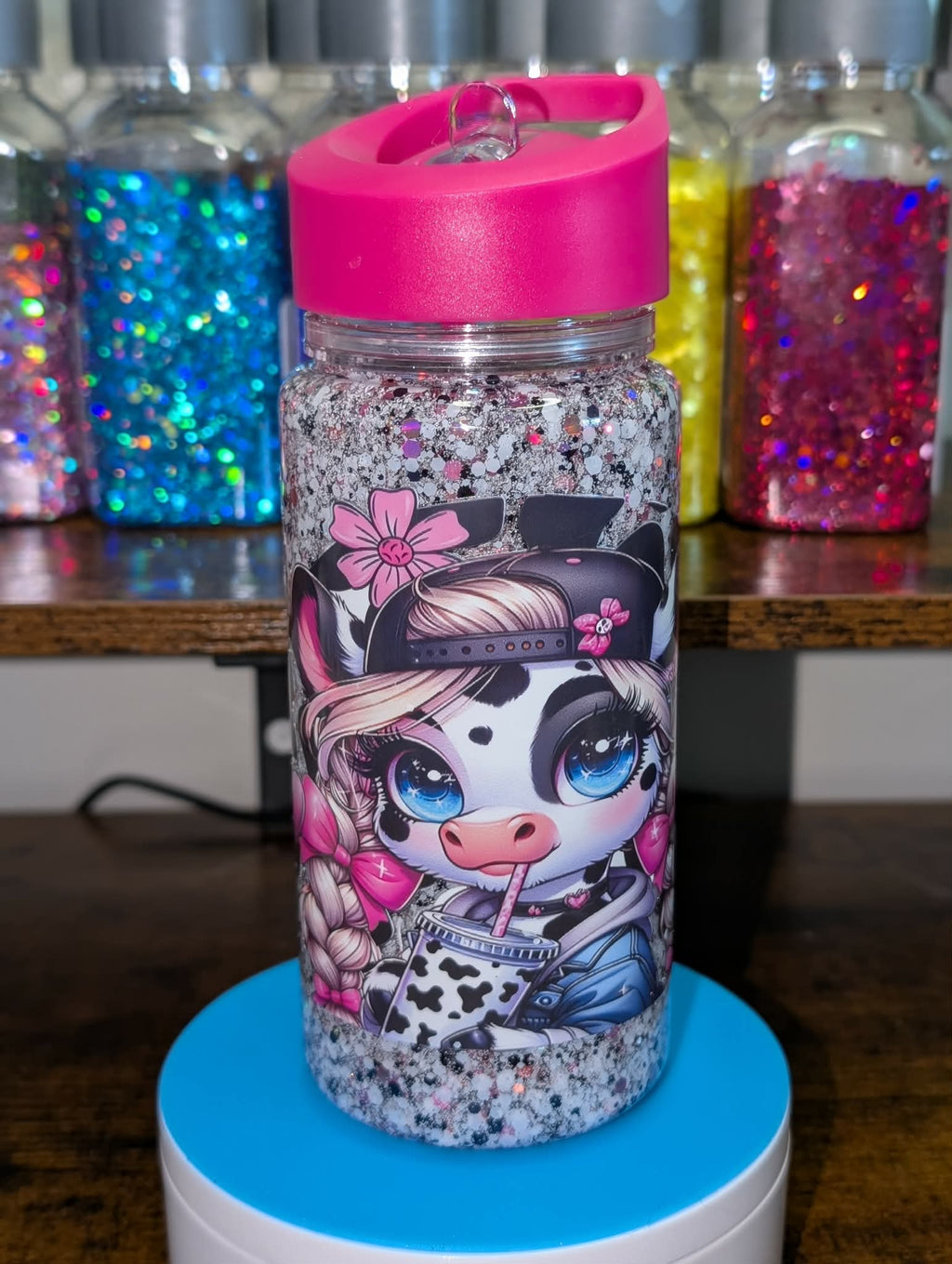 Glittery water bottle with cartoon character design on a blue stand, colorful jars in the background.