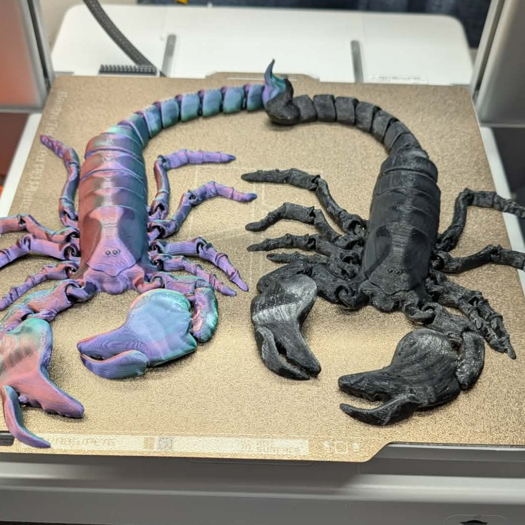Two 3D-printed scorpion models on a printing bed, one in black and one in multicolored.