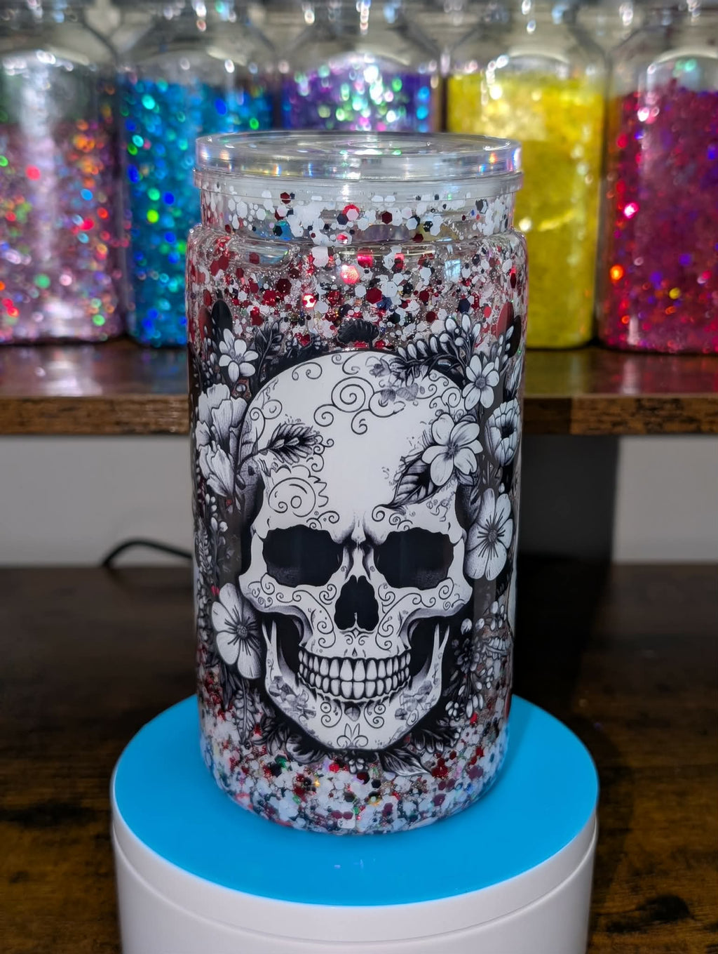 Tumbler with a skull design on a blue stand, surrounded by colorful glittery jars.